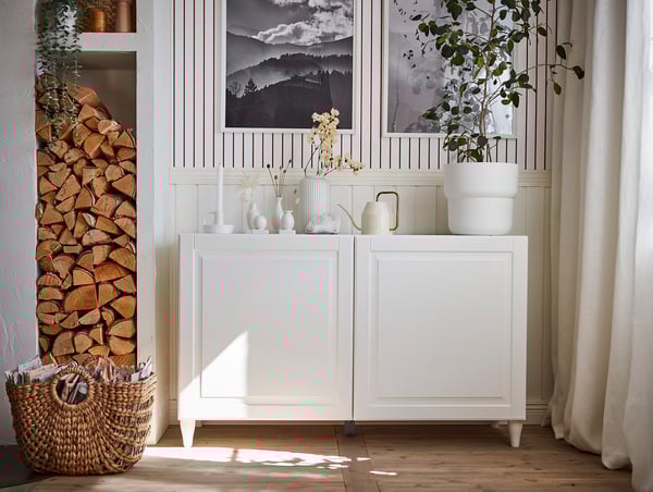 White BESTÅ sideboard with wooden floors, draped curtain, stacked logs, plants, vases, and framed artwork.