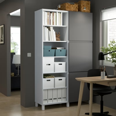 Modern grey BESTÅ storage unit, tall, with books, boxes, and decor on shelves. Includes desk and chair, well-lit room.