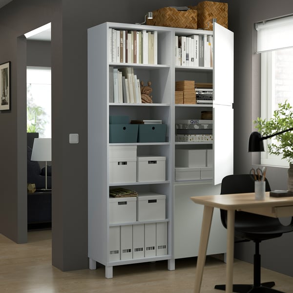 White BESTÅ storage unit with adjustable shelves, showcasing books and boxes, beside a wooden desk and black chair in a home office setting.