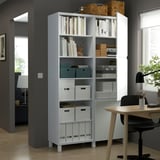White BESTÅ storage unit with adjustable shelves, showcasing books and boxes, beside a wooden desk and black chair in a home office setting.