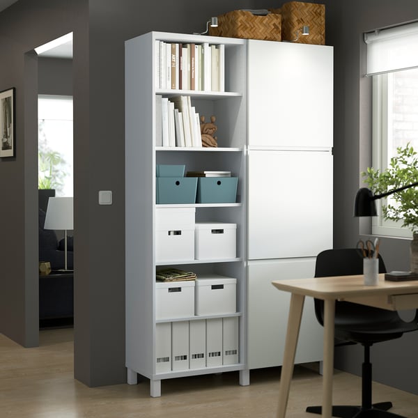 White BESTÅ storage unit in home office, holding books and boxes. Tall, fits room corner.