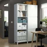 White BESTÅ storage unit in home office, holding books and boxes. Tall, fits room corner.