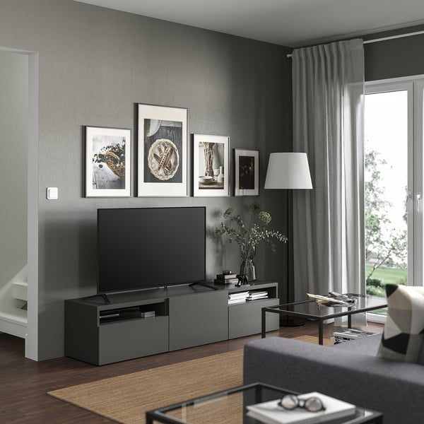 Modern living room with a BESTÅ tv bench in grey. It a flat-screen tv, framed art, grey curtains, and a glass coffee table on a beige rug.