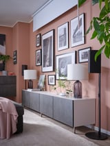 Modern living room with peach walls, featuring a grey BESTÅ tv unit with black lamps and plants. Various framed photos decorate the wall above.