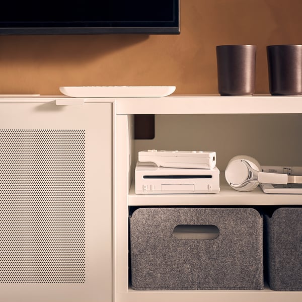 A white BESTÅ tv bench with adjustable shelves, housing a white gaming console, headset, and gray storage boxes, with a flat-screen tv above it.