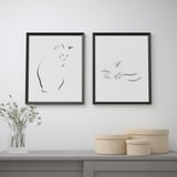 Two black-framed cat artwork pieces hang above a table with a vase of flowers and woven baskets.