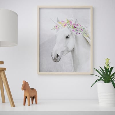 Framed artwork of a horse with a floral crown sits on a white shelf. A wooden horse figurine and a plant are beside it.