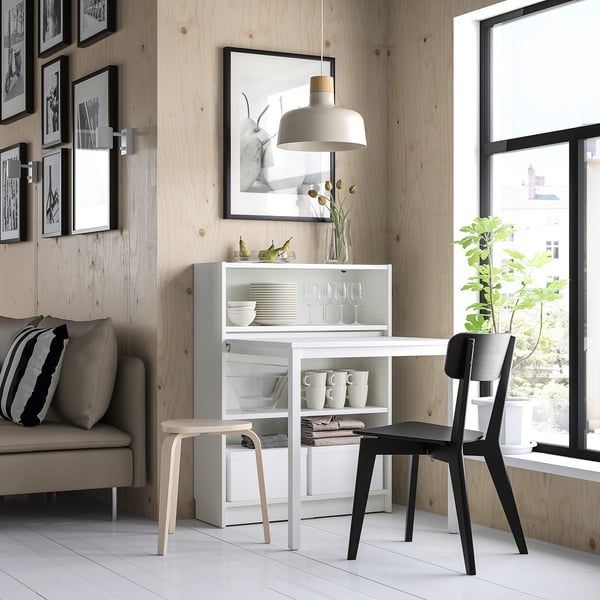 Modern dining area with white BILLY bookcase desk, black chair, and grey sofa. Wooden wall and large window.