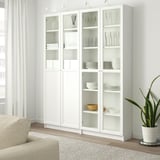 White BILLY bookcase, tall and narrow, four glass doors. Shelves hold dishes, books, jars. Plant and beige rug nearby. Tall in corner.