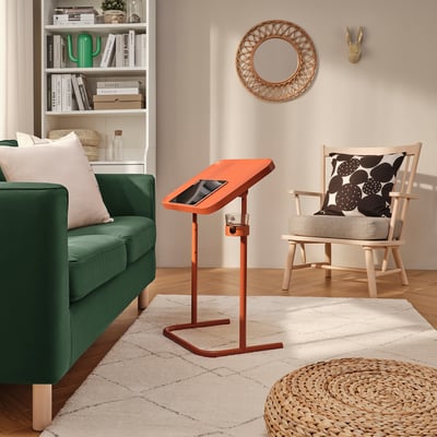 Bright, orange BJÖRKÅSEN laptop stand in cozy room. Its sleek, adjustable design perfectly fits the space between green couch and wooden chair.