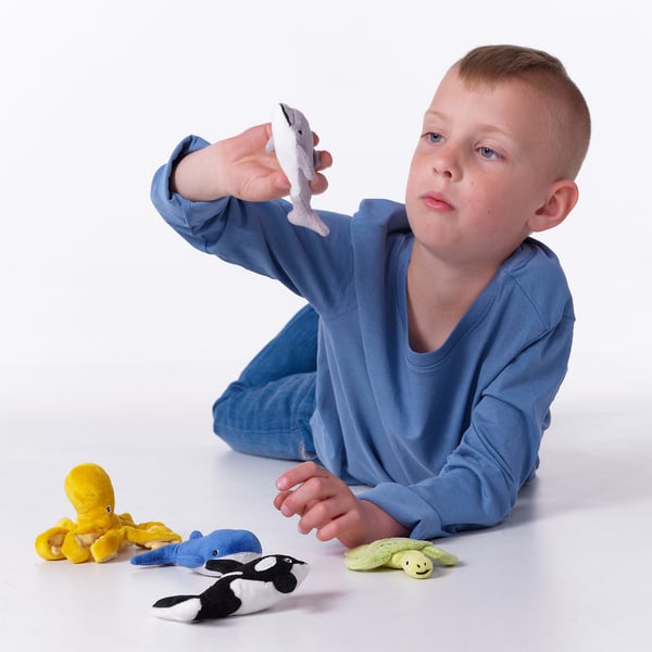 A boy plays with BLÅVINGAD soft sea creatures, holding one, surrounded by 4 more toys.