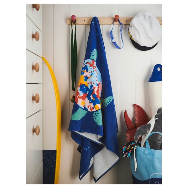 A colorful turtle-patterned blue bath towel hangs on a wooden hook next to a white wall and drawers with wooden knobs.