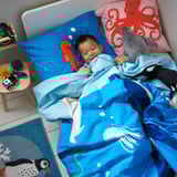 A toddler sleeps on a blue whale-patterned BLÅVINGAD duvet under a red octopus pillow, surrounded by ocean-themed decor.
