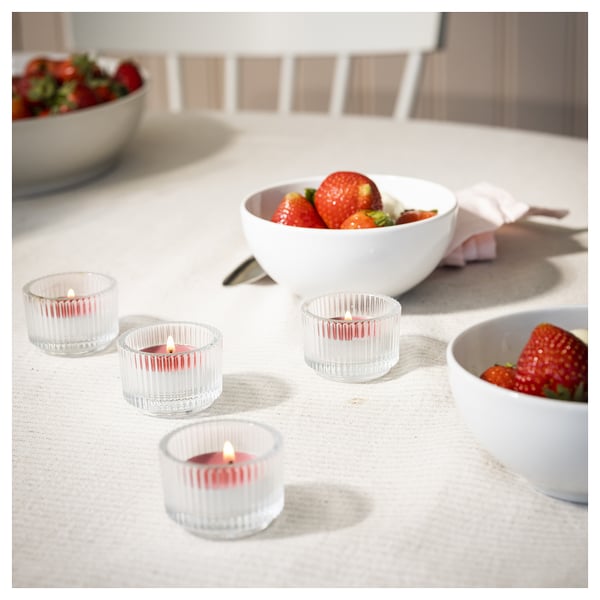 Four red striped glass candle cups with strawberry scent on a table with white bowls filled with strawberries.