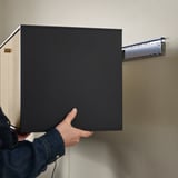 Person attaches modern black wall shelf with thin lines. Shelf has bi-fold oak veneer doors, enhancing storage and display.