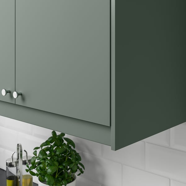 Green kitchen cabinets with modern handles. Underneath, a potted plant sits on a white tiled backsplash.