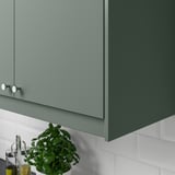 Green kitchen cabinets with modern handles. Underneath, a potted plant sits on a white tiled backsplash.