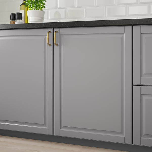 Modern grey kitchen cabinet with gold handles, set against a white tile backsplash and dark countertop.