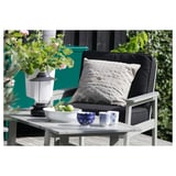 Modern outdoor setting with white wood chair, dark cushion, grey pillow, teal wall, green plants, and table with fruit and cups.