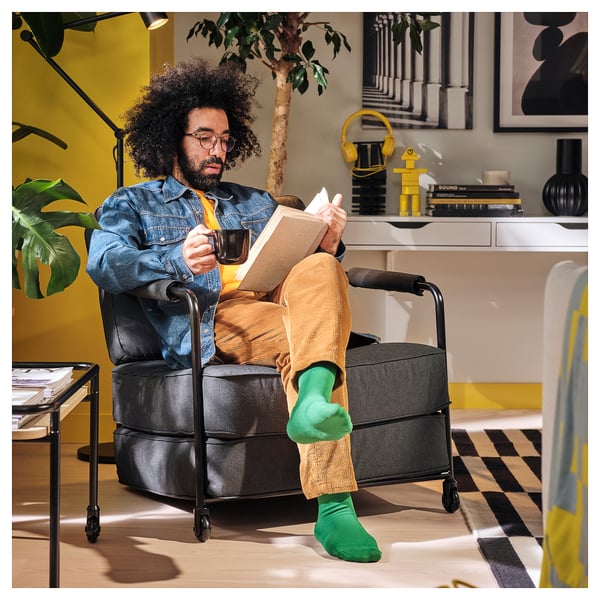 Man in denim jacket sits in versatile BRÄNNBOLL lounge chair, holding coffee and reading, showcasing its comfort and mobility.