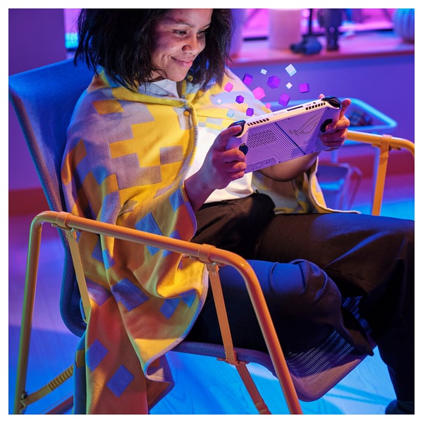 A person sits on a colorful chair, wrapped in a cozy pixel-patterned BRÄNNBOLL throw blanket, holding a gaming console.