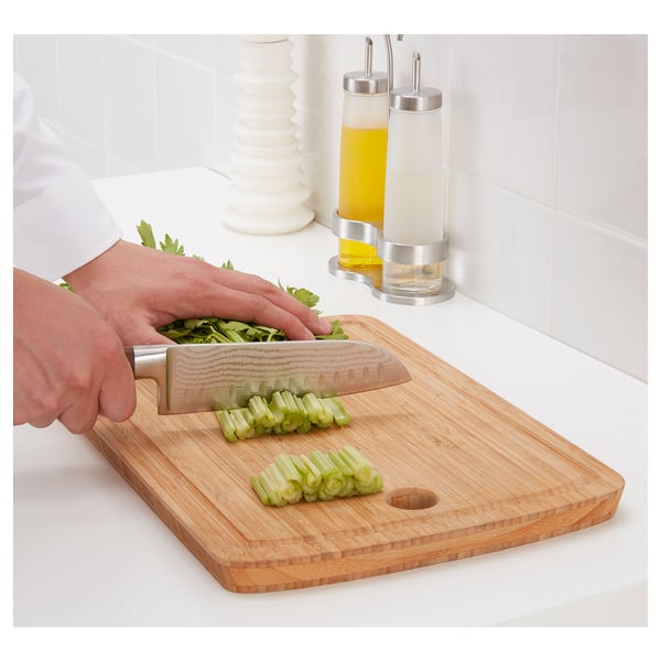 A person chops green vegetables on a bamboo cutting board using a stainless steel knife. The knife has a damascus steel blade.