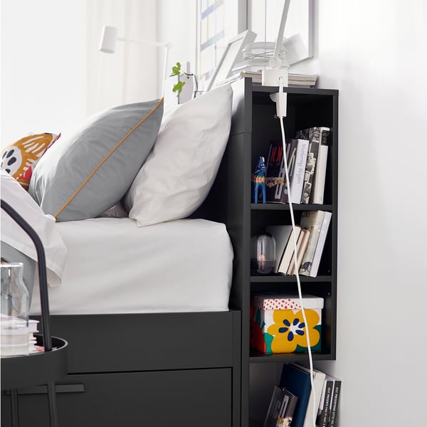 Modern bed with gray storage drawers and white bedding. Decorative pillows on a bed, books, and a lamp on a shelf beside it.