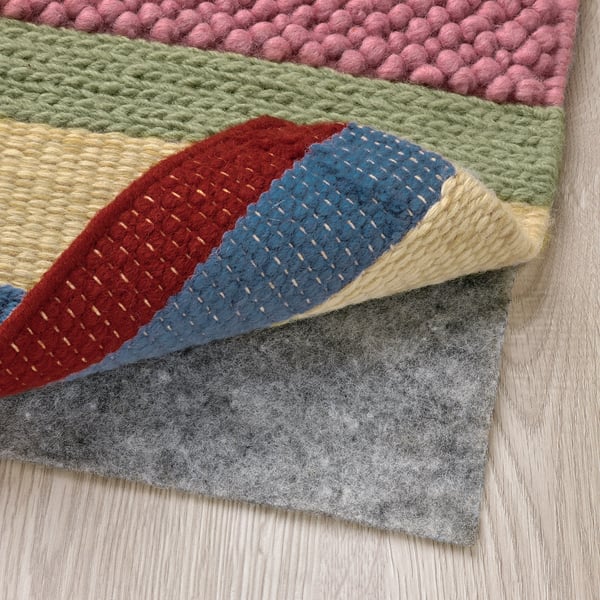 Bright, multicolored woven rug with varying textures, laid over a grey non-slip pad on wooden floor, showcasing its handcrafted quality.