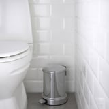 Silver trash pedal bin in bathroom beside white toilet with closing lid.