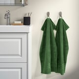 Two green BROKGLIM towels hang on hooks beside a bathroom sink, larger than toothbrush holder.