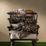 BRUDKRUSBÄR pillows: stacked black floral cushions on chair, larger below, smaller atop, vibrant flower designs.