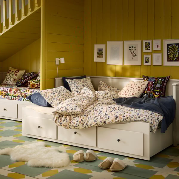 Bright yellow room with a white BRUDKRUSBÄR bed, floral bedding, colorful pillows, slippers, and wall art.