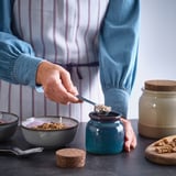 Person scooping granola from blue ceramic jar onto bowl. Jars and bowls displayed on a dark surface.