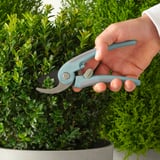 Hand holds light blue BRYTBÖNA pruners, cutting green bush; silicone grip and stainless steel blades evident.