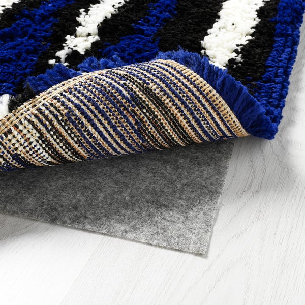 Colorful, fluffy rug with black, blue, and white patterns, rolled up on a grey nonslip mat on a wooden floor.
