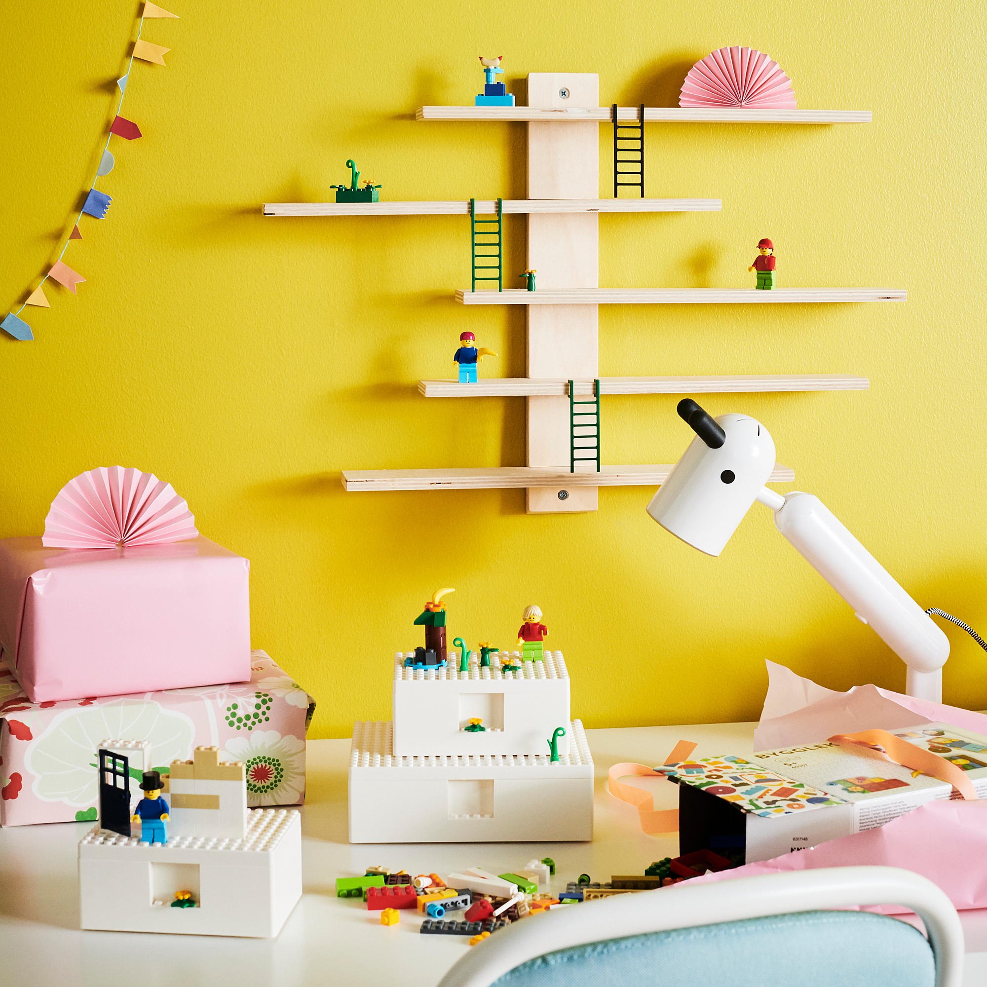 Kids play area with yellow wall, multilevel BYGGLEK toy set, lego figures, desk with more legos, lamp.