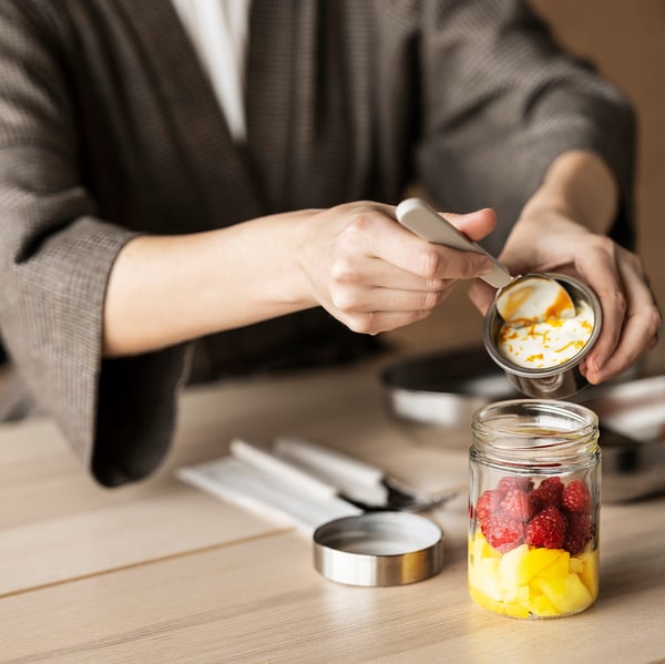 A person makes a fruit and yogurt parfait in a DAGKLAR jar with a separator on a wooden table among kitchen tools.
