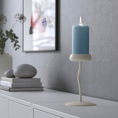 A lit teal blue DAGLIGEN candle, 6 tall, sits on unique white holder atop drawer, near books, rocks, and plant.