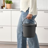 Person holding a gray bin with a handle, ideal for small waste like metal or batteries. The bin a removable lid for hiding waste.