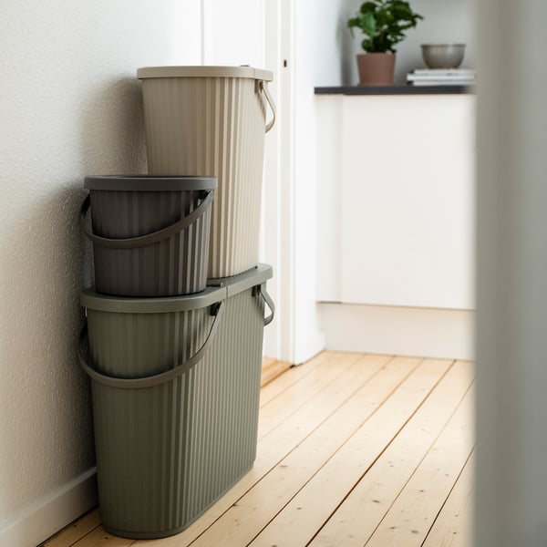 Three stacked DAMMÄNG bins in shades of beige and gray, rectangular with handles, neatly fitted against a wall in a modern, minimalist room.