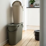 Three stacked DAMMÄNG bins in a corner, the top two beige, the bottom one dark green, with a smaller black bin beside them.