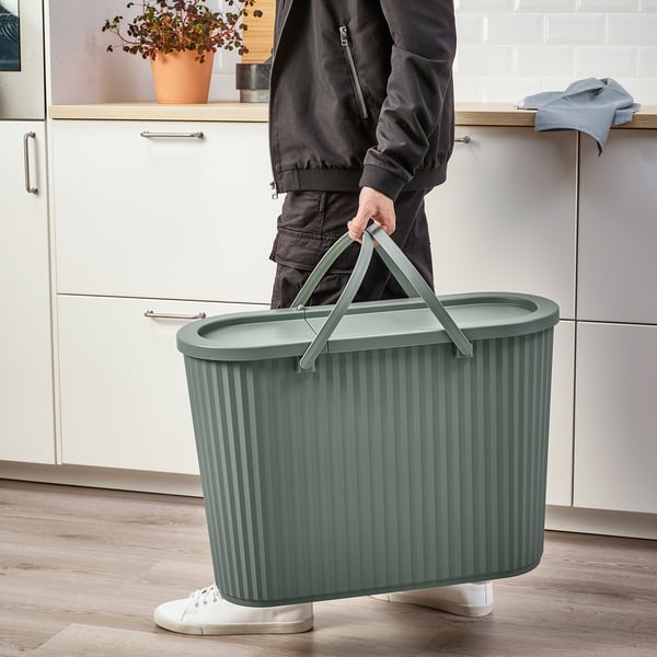 Person holding green waste bin with handle up, ready for transport. Lid hides contents, fits narrow spaces.