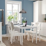Small white dining set with table and chairs, featuring black accents, set against blue wall, complemented by natural light and minimalistic decor.