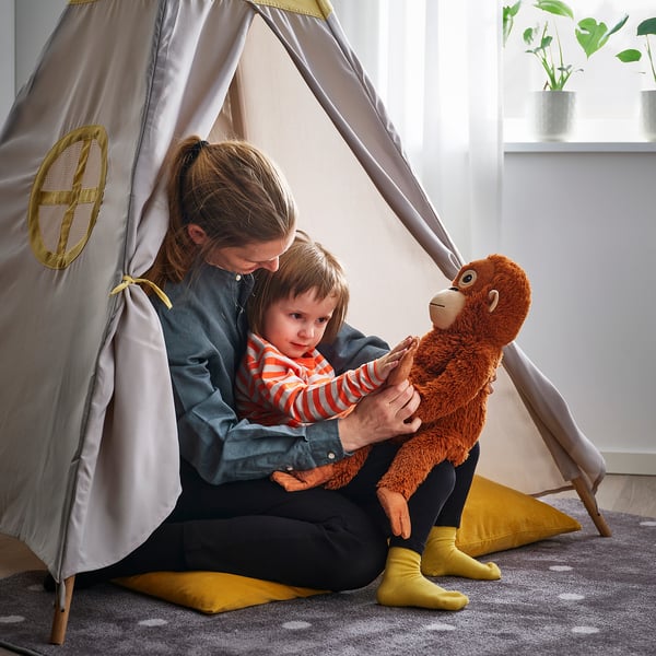 Woman & child play in tent. Child in pajamas holds toy, surrounded by pillows. Plant seen through window.