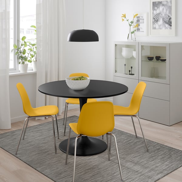 Bright dining area with black round DOCKSTA table, four yellow chairs, black pendant light, white curtains, grey rug, and potted plant.