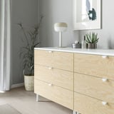 Wooden dresser with light knobs and handles; a simple, modern design; has three drawers, each with a smooth, rounded grip handle; sits on white legs.