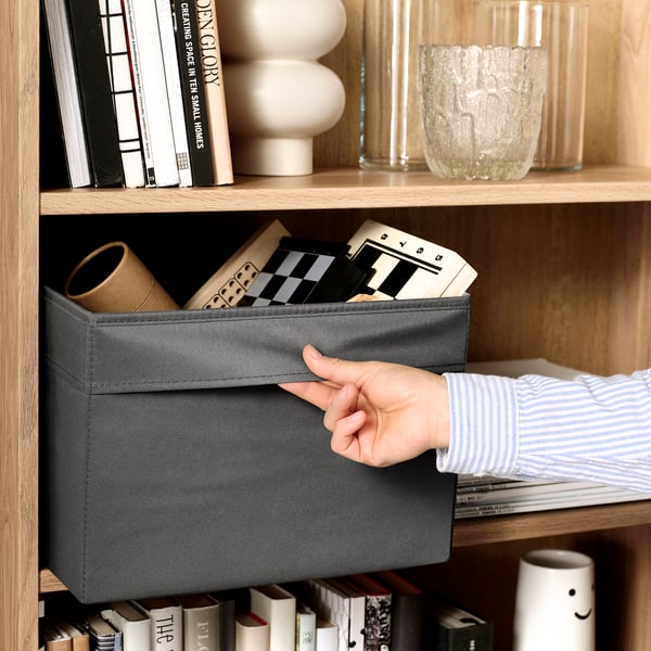 Hand reaches into grey DRÖNA storage box on wooden shelf, surrounded by books and decorative items.