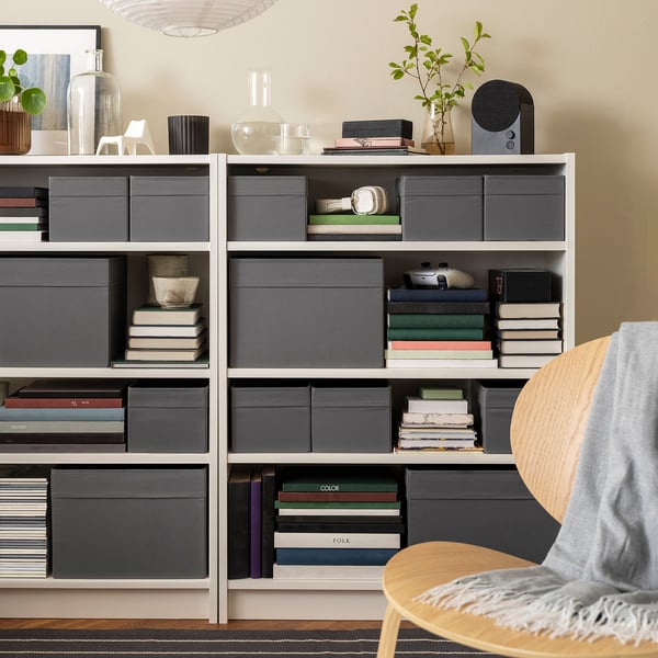 Grey DRÖNA boxes on shelves store books, bowls, plants.