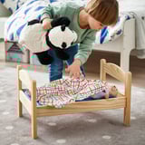 A child tucks a large stuffed panda into a wooden toy bed with a colorful blanket, promoting pretend play.