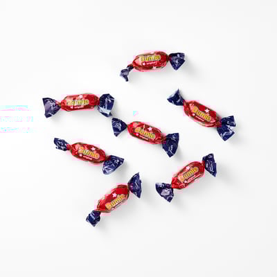 Eight red-wrapped DUMLE candies with blue ends are scattered on a white surface. DUMLE smooth chocolate and soft chewy toffee filling.